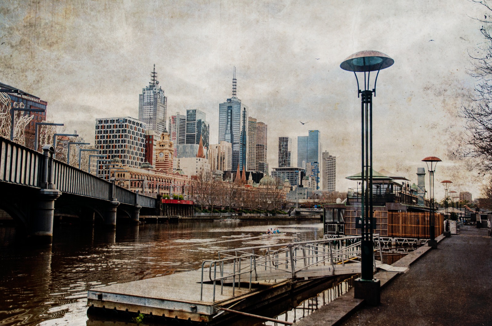 A riverside city view with landing pylons, layered buildings, and a muted skyline beyond the water.