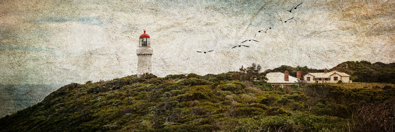 A white lighthouse with a red cap standing above low coastal vegetation and cottages on a windswept rise.