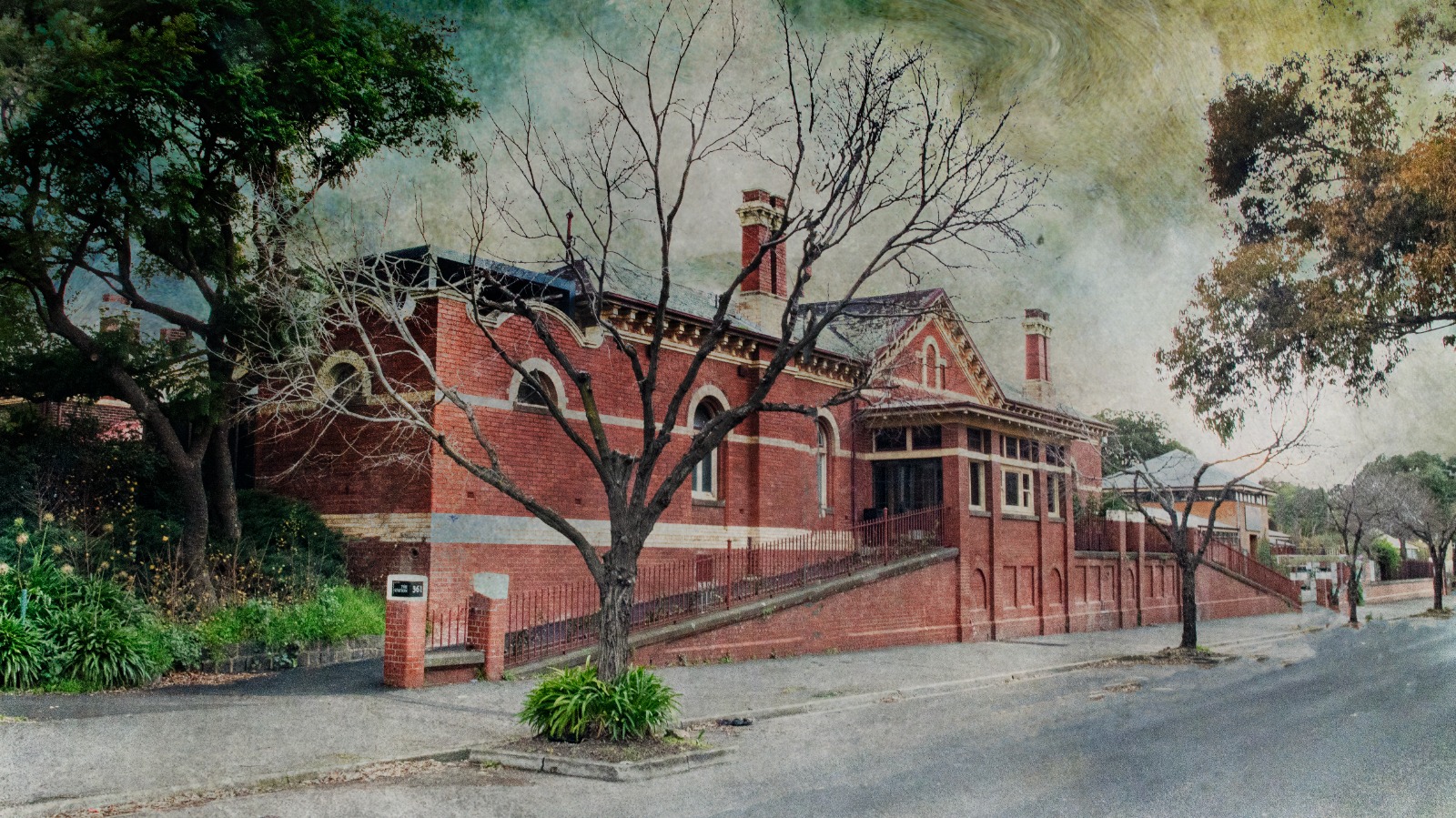A red brick heritage building with arched windows and chimneys facing a quiet street lined with trees.