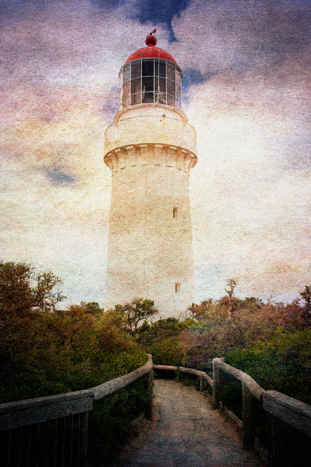 A white lighthouse with a red cap rising above a timber path and dense coastal planting.