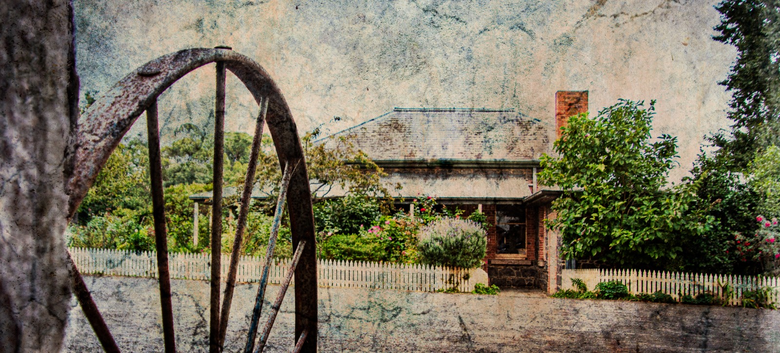 A rural homestead and picket fence seen beyond a rusted wagon wheel and garden growth.